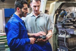 Two men in front of a large machine look at a tablet together