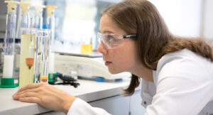 A woman in the laboratory wears a white coat and his protective goggles. She looks at test tubes